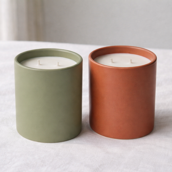Two candles in ceramic containers, one green and one terracotta, on a white surface.
