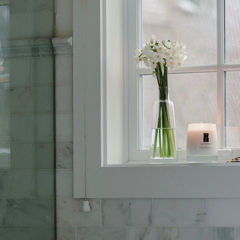 Luisa Terra Classic Candle in a sunlit bathroom with marble tile, white flowers, and an air of effortless Mediterranean elegance.